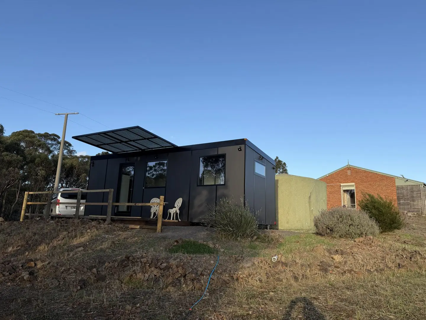 A charcoal MicroHome with a raised solar awning and a small deck, sited on a spare rural block.